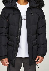 Parka negra acolchada con capucha de pelo sintético, cierres de cremallera y botón a presión, múltiples bolsillos y una etiqueta con el logotipo junto a la cremallera.