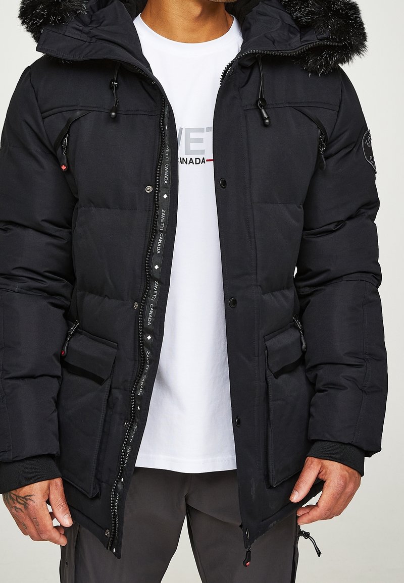 Parka negra acolchada con capucha de pelo sintético, cierres de cremallera y botón a presión, múltiples bolsillos y una etiqueta con el logotipo junto a la cremallera.
