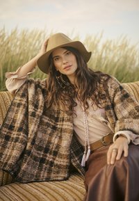 Stickad poncho i brunt och tan-rutat mönster med fransade kanter. Modellen bär en beige blus, brunt läderkjol och en tan bredbrättad hatt.