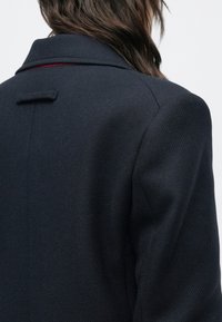 Marineblauwe jas van een gestructureerde stof, met een kraag met inkeping en een subtiel rood accent op de naden van de kraag, met minimalistische designelementen.