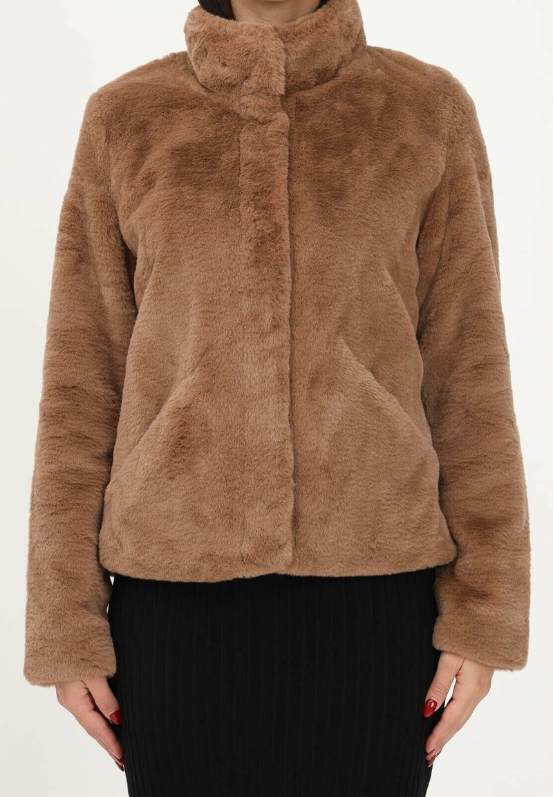 Personne portant une veste marron, duveteuse, à manches longues avec un col haut et une jupe noire côtelée, debout devant un fond uni.