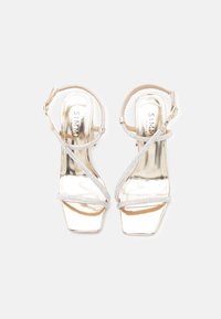 Χρυσά σανδάλια με λουράκια και ψηλό τακούνι, διακοσμημένα με κρυστάλλους, τετράγωνη μύτη και λαμπερό μεταλλικό φινίρισμα. Περιλαμβάνει ρυθμιζόμενο λουράκι στον αστράγαλο.