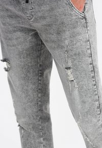 Pantalon en denim gris avec une coupe slim, des détails usés et des bords effilochés. Inclut des poches et une fermeture éclair.