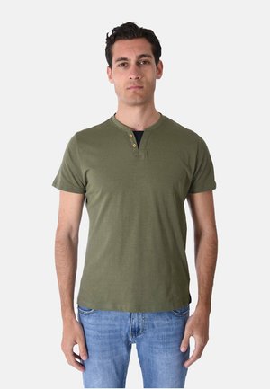 Uomo con capelli scuri che indossa una maglietta henley a maniche corte verde oliva e jeans blu, in piedi davanti a uno sfondo chiaro e uniforme.