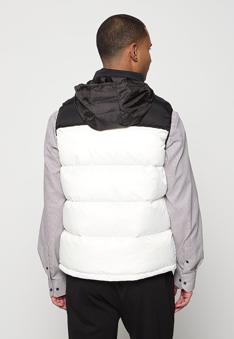 Gilet sans manches en duvet noir et blanc avec capuche, présentant des sections matelassées et une texture lisse, porté par-dessus une chemise à carreaux.