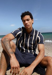 Homme tatoué portant une chemise à rayures bleu marine et blanche ainsi qu'un short bleu marine, assis sur une plage de sable avec des vagues de l'océan en arrière-plan.