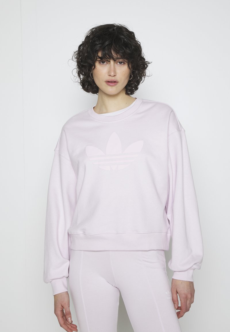 Gaiši rozā džemperis ar apaļu dekoltē, pārspīlētiem piedurknēm un tonālajām adidas emblēmām uz krūtīm. Mīksta auduma un brīvs griezums.