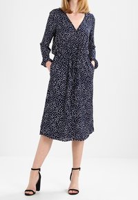 Abito blu navy con pois bianchi, scollatura a V, maniche lunghe, vita con cordoncino e tasche laterali, abbinato a tacchi neri con cinturino alla caviglia.
