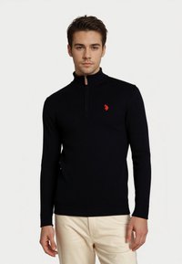 Junger Mann, der einen schwarzen langärmligen Half-Zip-Pullover mit einem roten Logo auf der Brust und hellbeigen Hosen trägt, vor einem schlichten Hintergrund.