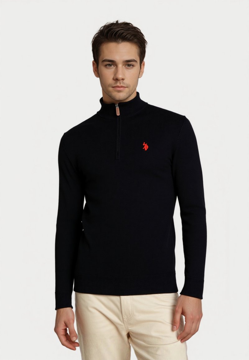 Junger Mann, der einen schwarzen langärmligen Half-Zip-Pullover mit einem roten Logo auf der Brust und hellbeigen Hosen trägt, vor einem schlichten Hintergrund.