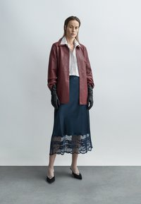 Rode leren jas, wit gestreept shirt, marineblauwe rok met kanten zoom, zwarte handschoenen en zwarte schoenen met puntige neus. Betonnen achtergrond.