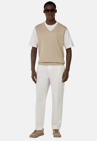 Gilet en tricot beige sur un t-shirt blanc, associé à un pantalon blanc et des baskets beiges. Le gilet possède un col en V et une texture côtelée.