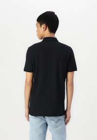 Polo preto com colarinho, mangas curtas e bainha reta, feito de um tecido suave. Usado com jeans de tom azul claro com costura nos bolsos traseiros.