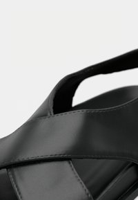 Η μαύρη σανδάλι διαθέτει σχεδιασμό με σταυρωτά λουριά από λείο υλικό. Στρογγυλεμένο σχήμα με ορατές λεπτομέρειες ραφών και ανοιχτή φτέρνα για καλύτερο αερισμό.