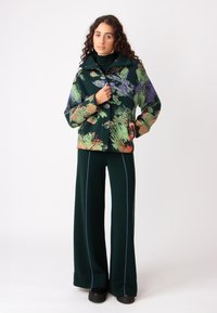 Zelena jakna s potiskom, ki vključuje vijolične, oranžne in zelene cvetlične vzorec, z gumbi spredaj, v kombinaciji s temno zelenimi širokimi hlačami z modrimi detajli.