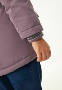 Veste matelassée violette pour enfant avec une finition texturée, présentant une manche à poignet sur une couche intérieure grise rayée. Une main saisissant l ourlet.