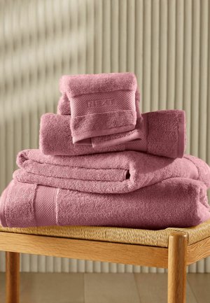 Pila di asciugamani rosa polvere piegati con bande testurizzate, appoggiati su un sedile intrecciato in legno contro uno sfondo beige rigato.