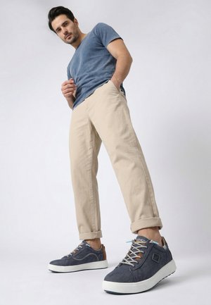 Mann in blauem T-Shirt und beiger Hose, der blaue Freizeitsneaker mit weißen Sohlen trägt und vor einfachem hellem Hintergrund steht.