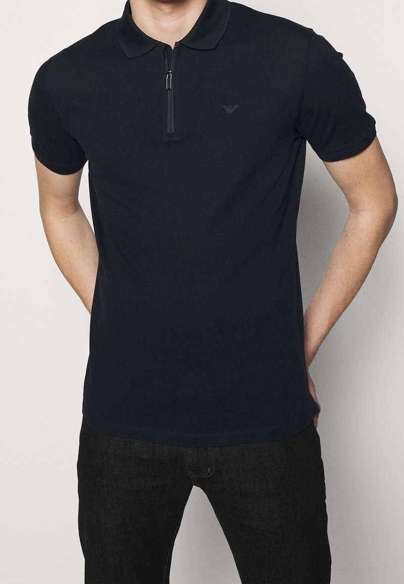 Černé polo triko s krátkým zipem, krátkými rukávy a jemným logem na hrudi; vyrobené z hladkého materiálu s přiléhavým střihem.