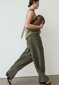 Kvinde iført olivengrønne vide bukser, ærmeløs striktop med bindebånd i halsen, holder stor vævet brun håndtaske, iført sorte sandaler.