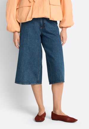 Donna che indossa pantaloncini di jeans blu a gamba larga di lunghezza media, una camicetta pesca con maniche a sbuffo e ballerine rosse intrecciate con punta.