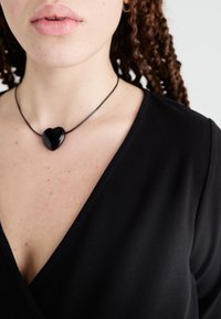 Haut noir à col en V avec une texture lisse, associé à un pendentif en forme de cœur noir sur un collier en cordon fin, reposant sur la clavicule.