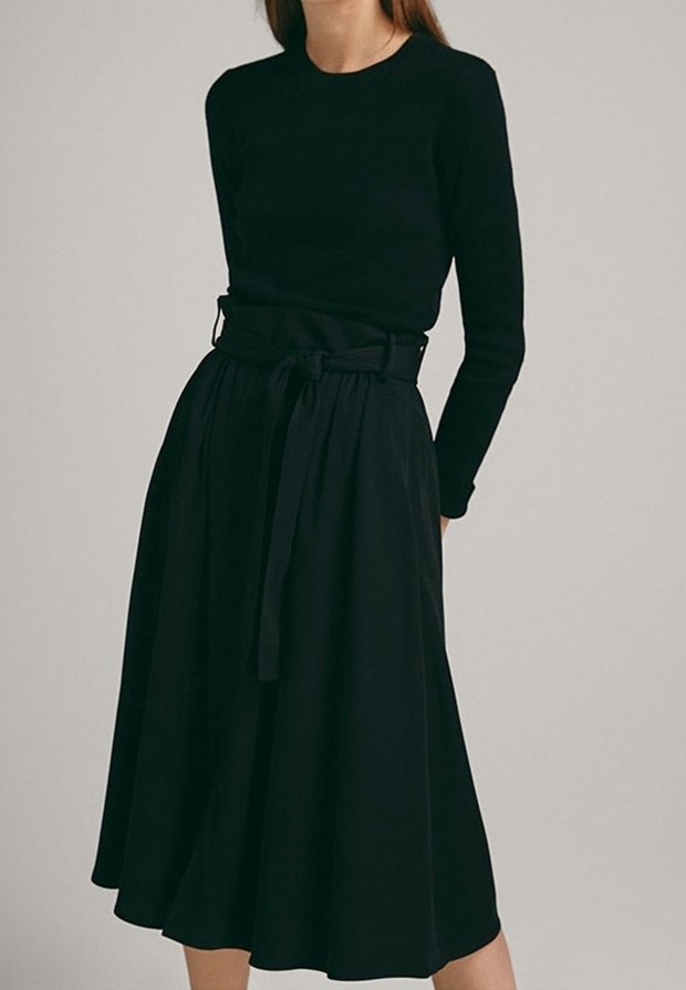 Femme portant un haut noir à manches longues et une jupe midi noire taille haute avec une ceinture nouée, debout devant un fond uni.