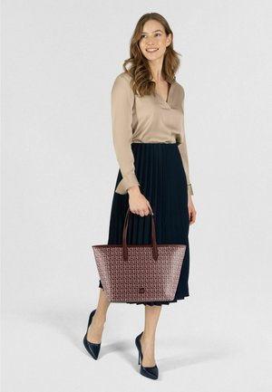 Bordeauxrote Tragetasche mit Logo-Muster, kombiniert mit einer beigen Bluse und einem plissierten schwarzen Rock. Die Tasche verfügt über braune Griffe und eine strukturierte Form.