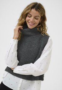 Sivi, rebrasti džemper s visokim ovratnikom i ležernim krojem, nosi se preko bijele košulje s ovratnikom koja ima suptilne otisnute naglaske. Mekana tekstura, skraćena dužina.