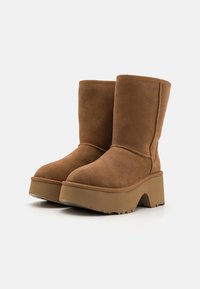 Botas de tornozelo em camurça castanha com solas de plataforma alta e bico redondo. Apresentam detalhes em costura e uma textura suave.