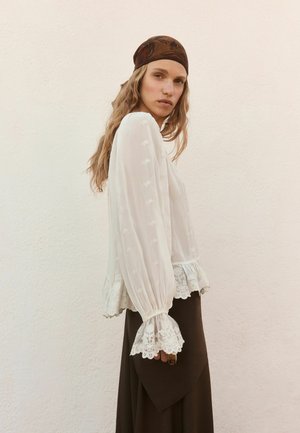 Kvinde iført hvid blondetrimmet bluse og mørkt skørt, med langt bølget hår dækket af et brunt mønstret tørklæde, stående sideværts.