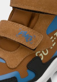 Primo piano di una tracolla di sneaker marrone con "PMG" in blu, accenti blu e neri, e del testo parziale "GULL" sul lato della scarpa.