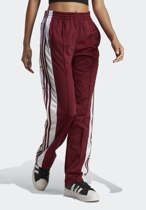 Maroon track pants with white side stripes, elastic waistband, and snap-button detail along the sides. Paired with black and white sneakers.