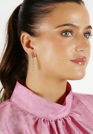 Guldtonade örhängen med länkade rektangulära former, prydda med klara stenar. Bärs av en modell i en pink, strukturerad blus med hög krage.