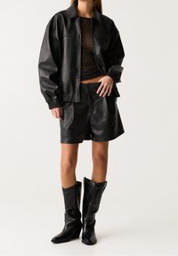 Veste en cuir noire avec poignets élastiques, haut noir transparent parsemé de points argentés, short en cuir assorti et bottes de cowboy noires montantes jusqu'aux genoux.