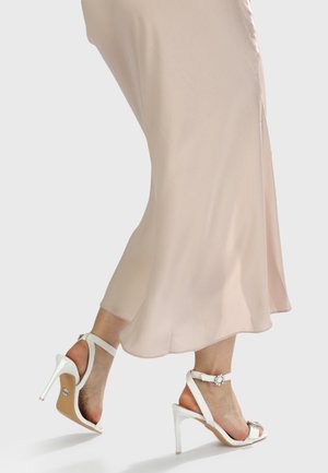 White high-heeled sandals with a jeweled accent and straps. Worn with a soft, flowing beige dress, showcasing the shoe's elegant design.