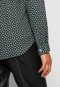 Gros plan sur la main d’une personne reposant le long de son corps, portant une chemise bleu marine à motifs avec de petits motifs blancs et verts et un pantalon en cuir noir.
