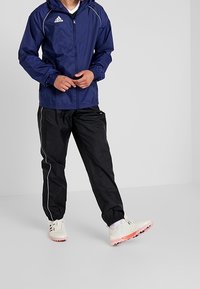 Veste légère bleue avec capuche, poignets élastiques et accents blancs ; associée à un pantalon noir avec des bandes blanches sur les côtés et des chaussures blanches.