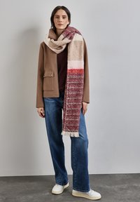 Sciarpa lavorata a maglia con strisce bordeaux, crema e bianche, caratterizzata da una finitura testurizzata. Indossata sopra una giacca marrone e abbinata a jeans blu.
