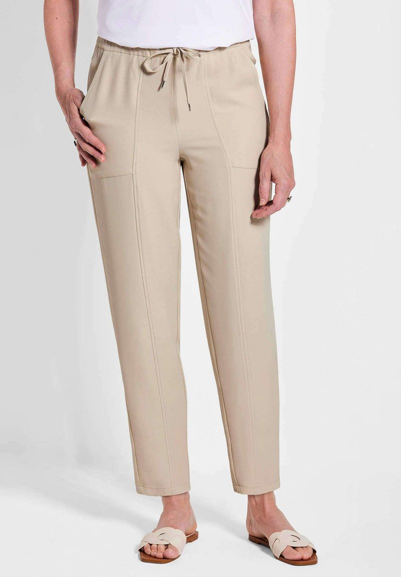 Beige instapbroek met een elastische tailleband, zijzakken, platte voornaden en een taps toelopende pijp. Gekleed met lichtgekleurde instapsandalen.