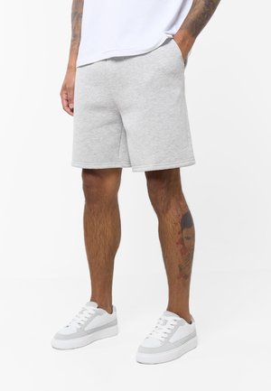 Man draagt lichtgrijze shorts, witte sneakers en een wit shirt, staat met handen in de zakken en toont een tatoeage op de linker kuit.