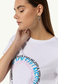 White cotton t-shirt featuring colorful text in a circular shape. Model wears silver hoop earrings and touches her neck. Smooth fabric texture.