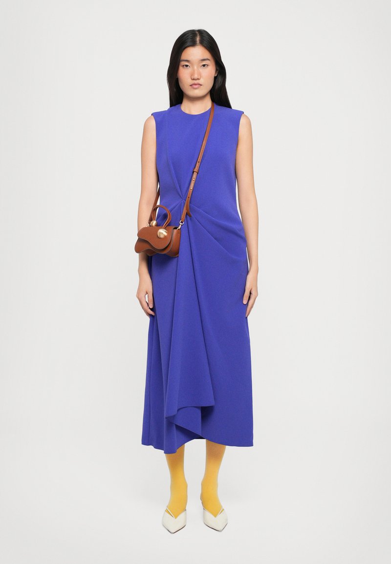 Une robe sans manches violette avec des détails froncés, associée à un sac bandoulière marron, des collants jaunes et des escarpins blancs à bouts pointus.