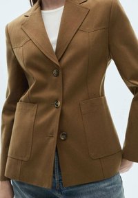 Femme portant un blazer marron sur mesure à trois boutons, avec de grandes poches avant, par-dessus un haut blanc et un jean bleu.