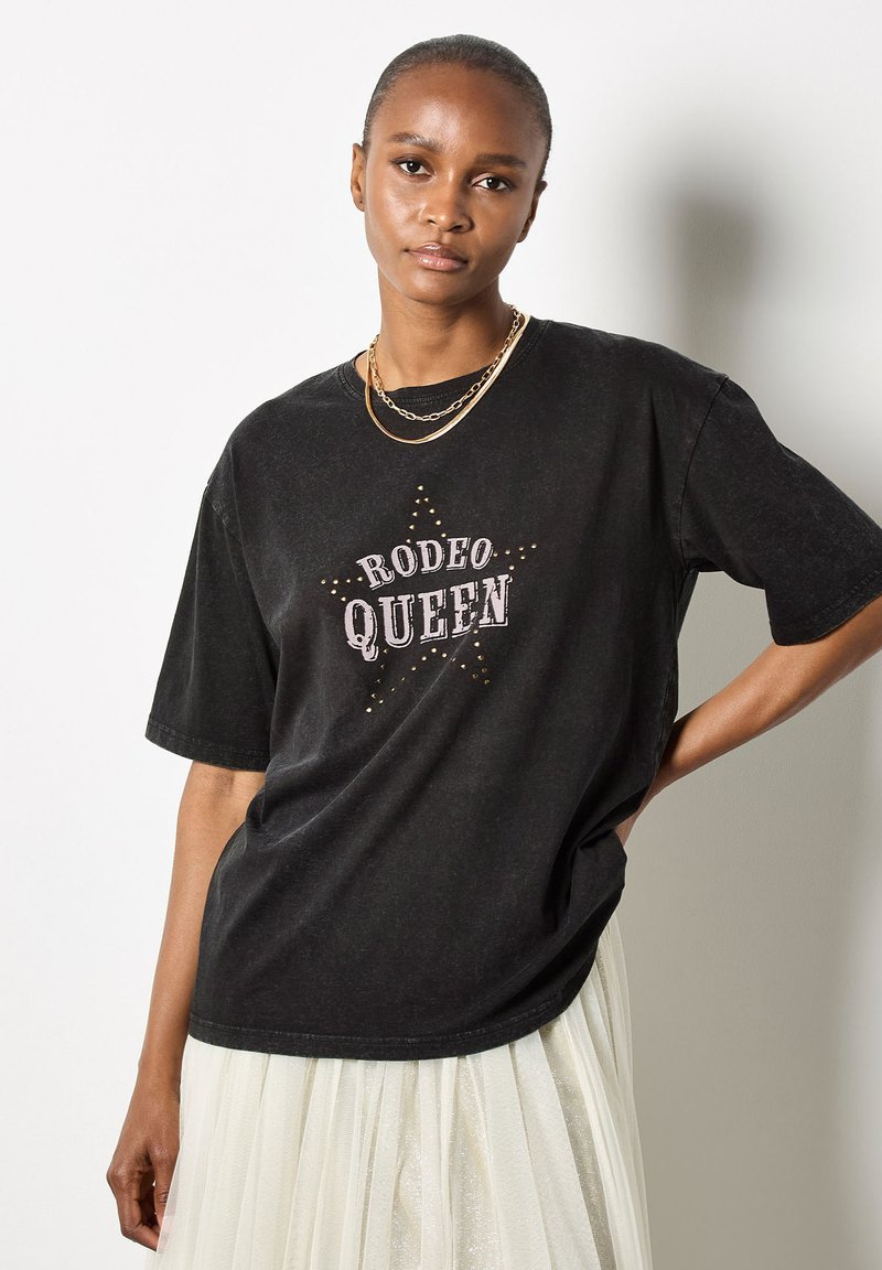 Zwarte oversized t-shirt van katoen met de tekst "RODEO QUEEN" en sterontwerp, voorzien van studs. Het model draagt gelaagde gouden kettingen.
