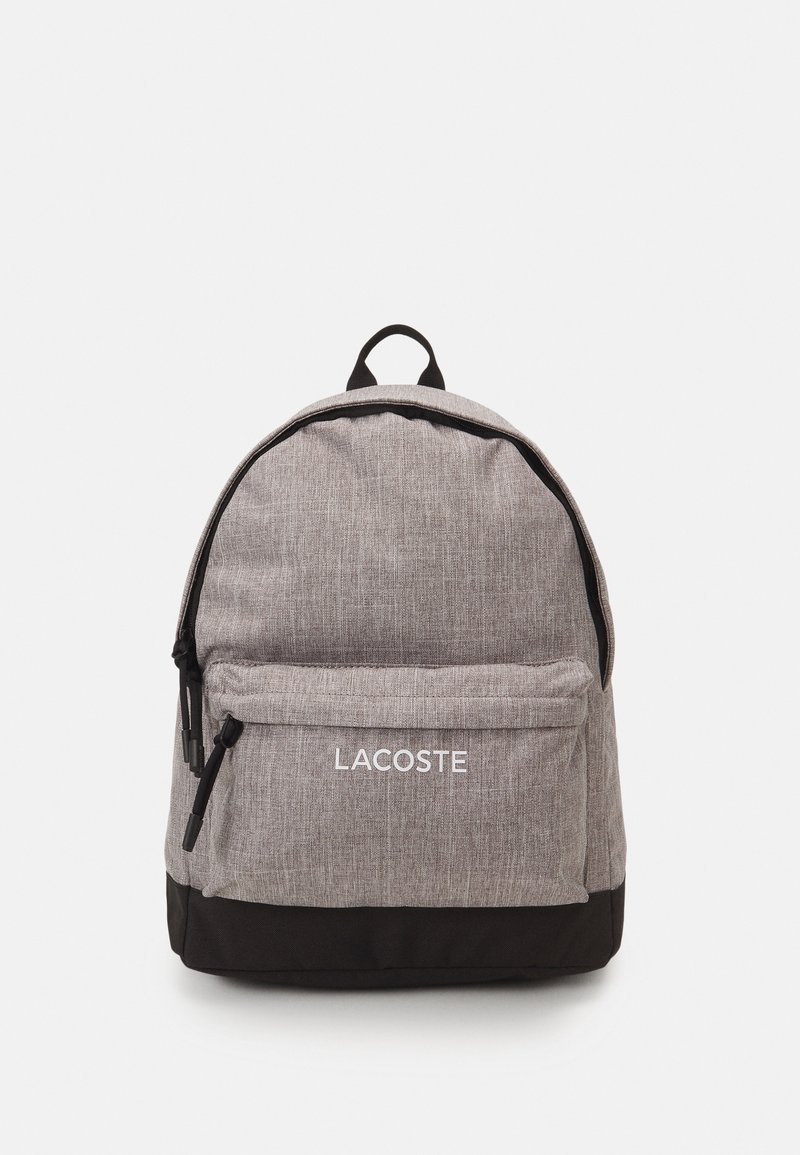 Sac à dos en tissu gris de forme arrondie, poche avant, fond noir et logo blanc "LACOSTE". Il dispose d'une poignée supérieure et de bretelles réglables.