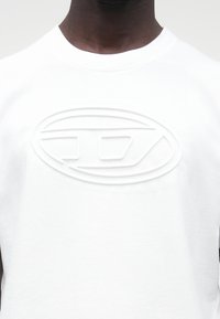 Artimas vaizdas asmens, dėvinčio baltą marškinius su pakilusia ovalo formos iškilusia emblema, esančia krūtinės centre.