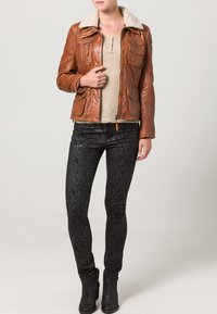 Bruine leren jas met een kraag van schapenvacht, vooraan met knopen, vier zakken aan de voorkant, gecombineerd met zwarte skinny jeans met patroon en enkellaarsjes.