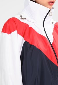 Femme portant un coupe-vent zippé à col montant avec des blocs de couleur blanc, rouge et bleu marine et un cordon de serrage argenté à la capuche.