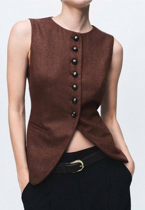 Ærmeløs brun vest med sorte knapper båret over sorte bukser med sort bælte og guld spænde, hænderne i lommerne, beskåret ved maven.